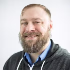 Smiling man with a full beard wearing a hoodie against a light background.
