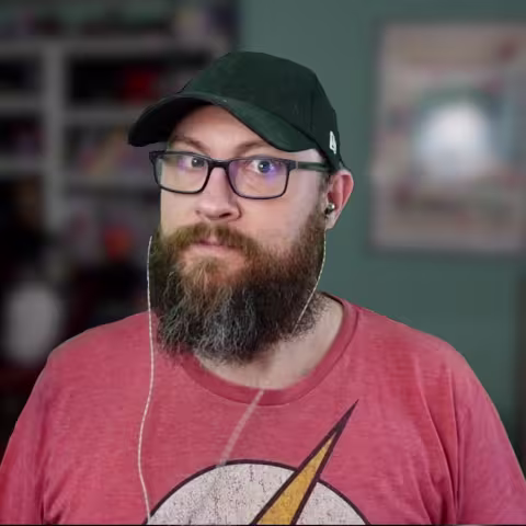 Man with glasses, a beard, and a black cap wearing a Flash logo shirt and earbuds indoors.