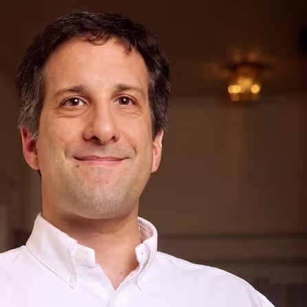 Smiling man wearing a white button-up shirt indoors with a softly blurred background.