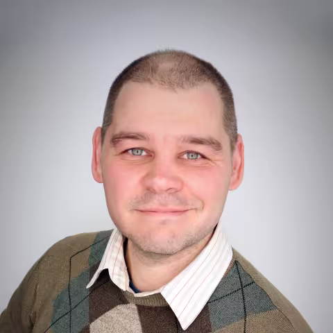 Smiling man with closely cropped hair wearing a collared shirt and patterned sweater against a light background.
