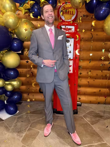 Man in a gray suit with a pink tie and loafers standing in front of festive balloons and a vintage gas pump backdrop.