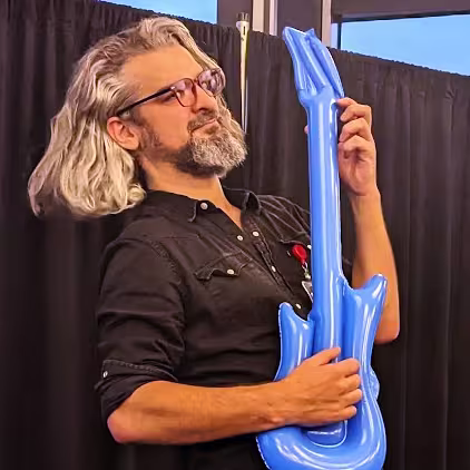 Person with long gray hair and glasses playfully holding a blue inflatable guitar.