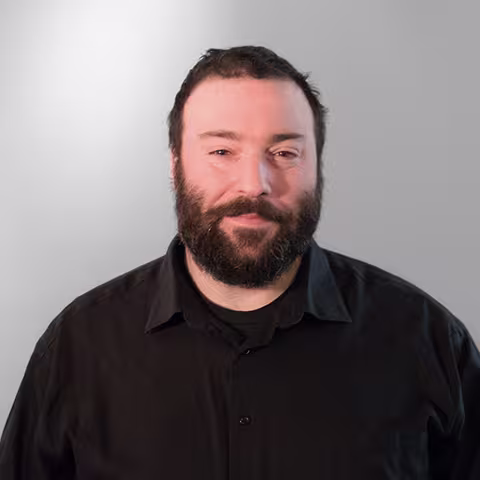 Man with a full beard wearing a black button-up shirt against a light background.