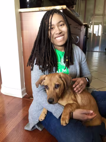 Smiling person with long dreadlocks sitting indoors while holding a brown puppy.