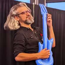 Person with long gray hair and glasses playfully holding a blue inflatable guitar.