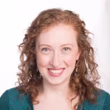 Smiling person with curly red hair against a light background.