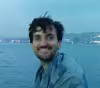 Smiling man with dark hair sitting near the water at dusk with city lights in the background.