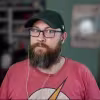Man with glasses, a beard, and a black cap wearing a Flash logo shirt and earbuds indoors.