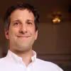 Smiling man wearing a white button-up shirt indoors with a softly blurred background.