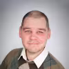 Smiling man with closely cropped hair wearing a collared shirt and patterned sweater against a light background.