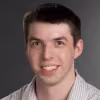 Smiling man with short dark hair wearing a light checkered shirt against a gray background.