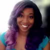 Smiling person with long wavy hair fading from dark to purple, wearing a teal top.